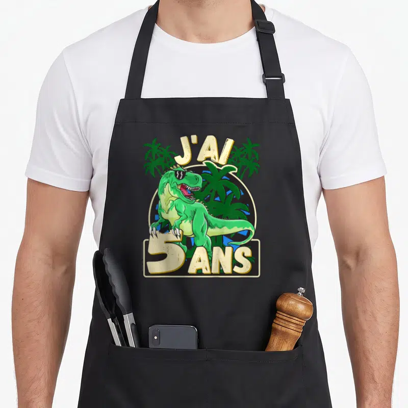 Tablier de cuisine noir avec un dinosaure vert portant des lunettes de soleil et le texte J'ai 5 ans entouré de palmiers.