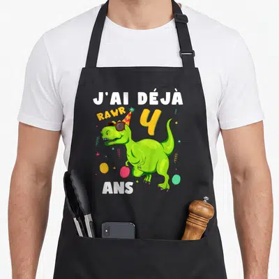 Tablier de cuisine noir avec dinosaure vert portant des lunettes de soleil et un chapeau de fête entouré de ballons.