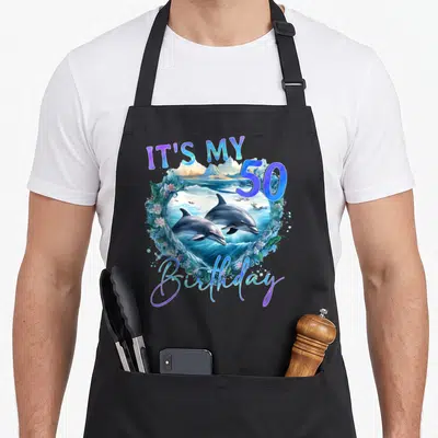 Tablier de cuisine noir avec illustration de dauphins, vagues et texte IT'S MY 50 anniversaire coloré.