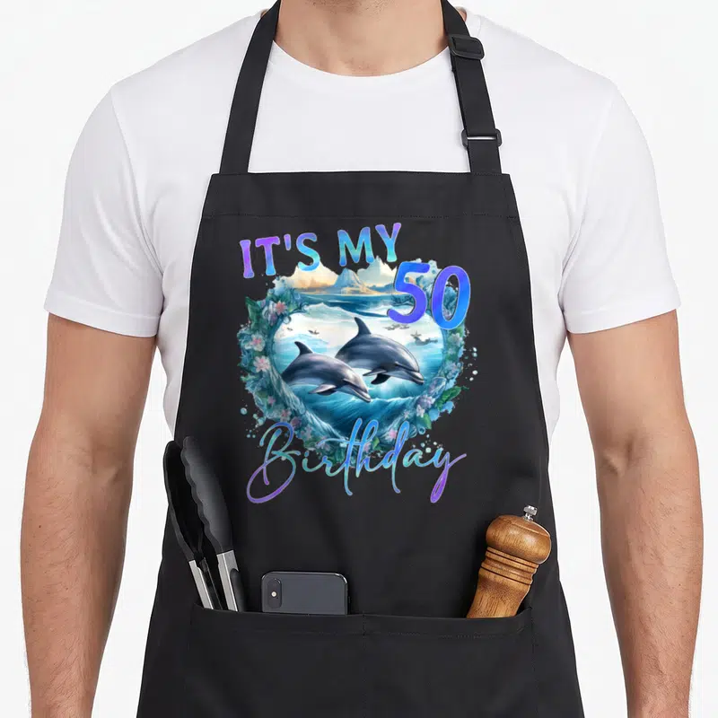 Tablier de cuisine noir avec illustration de dauphins, vagues et texte IT'S MY 50 anniversaire coloré.
