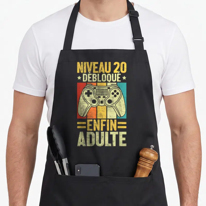 Tablier de cuisine noir avec illustration de manette de jeu vidéo et texte Niveau 20 Débloqué Enfin Adulte sur fond de bandes colorées.