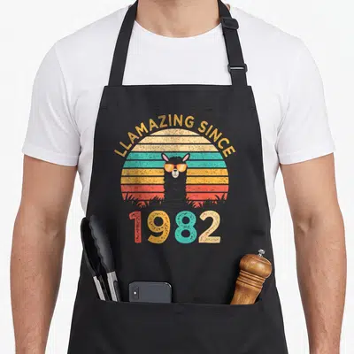 Tablier de cuisine noir avec un lama aux lunettes de soleil et le texte LLamazing Since 1982 en style rétro.