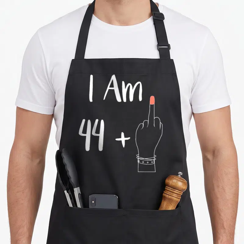 Tablier de cuisine noir avec l'inscription I Am 44 plus un doigt levé et un bracelet au poignet.