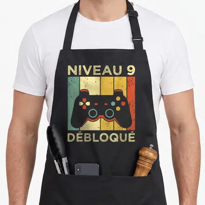 Tablier de cuisine noir avec manette de jeu colorée et texte Niveau 9 Débloqué en style vintage.