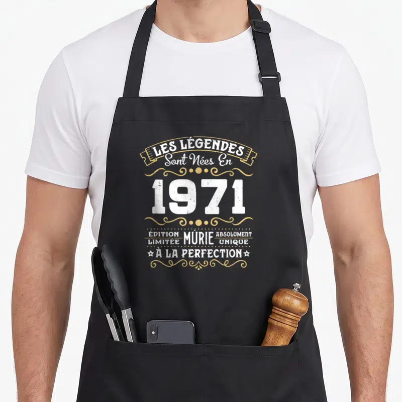 Tablier de cuisine noir avec le texte Les Légendes Sont Nées En 1971 en typographie vintage blanche et dorée.