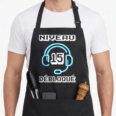 Tablier de cuisine noir avec illustration casque gaming bleu et texte Niveau 15 Débloqué en pixel art blanc.