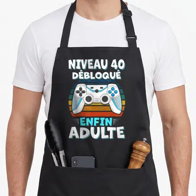 Tablier de cuisine noir avec une manette de jeu vidéo blanche et le texte Niveau 40 Débloqué Enfin Adulte en blanc et bleu cyan.