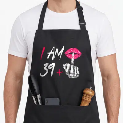 Tablier de cuisine noir avec l'inscription J'ai 39 ans et un majeur accompagnée d'une main squelettique rose.