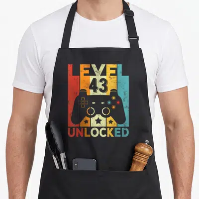 Tablier de cuisine noir avec illustration rétro d'un contrôleur de jeu et le texte Level 43 Unlocked en couleurs vives.