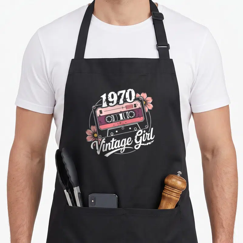Tablier de cuisine noir avec illustration de cassette rose et texte Vintage Girl 1970 entouré de fleurs.