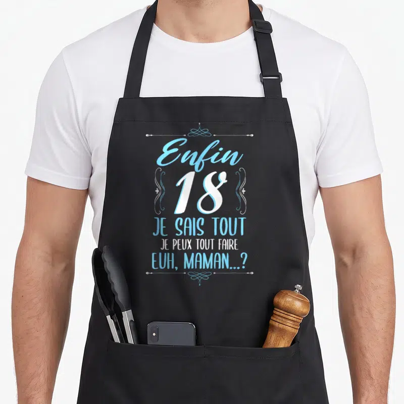 Tablier de cuisine noir avec l'inscription « Enfin 18 Je Sais Tout » en bleu et blanc entouré de volutes décoratives.