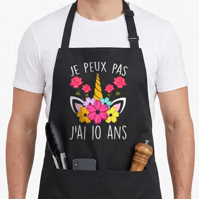 Tablier de cuisine noir avec corne de licorne dorée, roses roses et fleurs multicolores entourant des oreilles blanches stylisées.