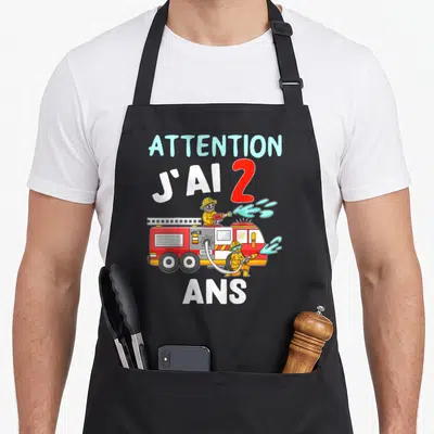 Tablier de cuisine noir avec une illustration de pompier, un camion rouge et le texte Attention j'ai 2 ans en lettres colorées.