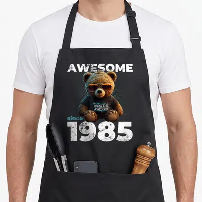 Tablier de cuisine noir avec un ours portant des lunettes de soleil et le texte « GÉNIAL depuis 1985 » imprimé au centre.