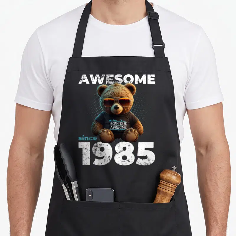 Tablier de cuisine noir avec un ours portant des lunettes de soleil et le texte « GÉNIAL depuis 1985 » imprimé au centre.
