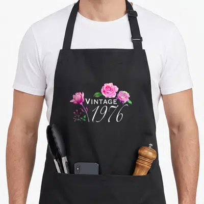 Tablier de cuisine noir avec trois roses roses et l'inscription Vintage 1976 en blanc au centre du motif floral.