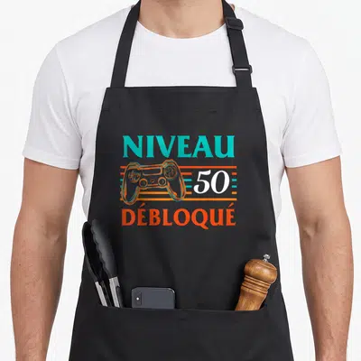 Tablier de cuisine noir avec l'inscription Niveau Débloqué 50 ans et une illustration de manette de jeu orange et turquoise.
