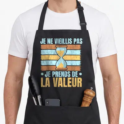 Tablier de cuisine noir avec un sablier coloré et le texte Je ne vieillis pas je prends de la valeur imprimé au centre.
