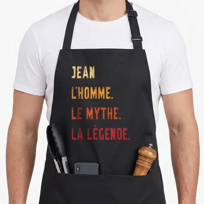 Tablier de cuisine noir avec le texte Jean l'homme le mythe la légende imprimé en lettres colorées dégradées.