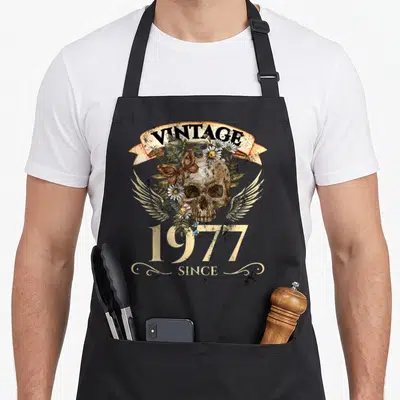 Tablier de cuisine noir avec une tête de mort vintage, des marguerites, un papillon et l'inscription 1977 since.