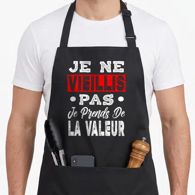 Tablier de cuisine noir avec le texte Je Ne Vieillis Pas Je Prends De La Valeur en typographie blanche et rouge.