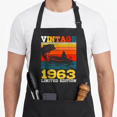 Tablier de cuisine noir avec motif vintage 1963 représentant un pêcheur en bateau et un poisson sautant sur des bandes colorées.