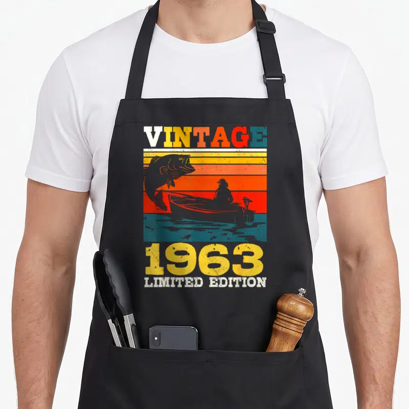 Tablier de cuisine noir avec motif vintage 1963 représentant un pêcheur en bateau et un poisson sautant sur des bandes colorées.