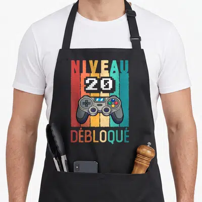 Tablier de cuisine noir avec illustration rétro d'une manette de jeu vidéo et texte Niveau 20 Débloqué en lettres colorées verticales.