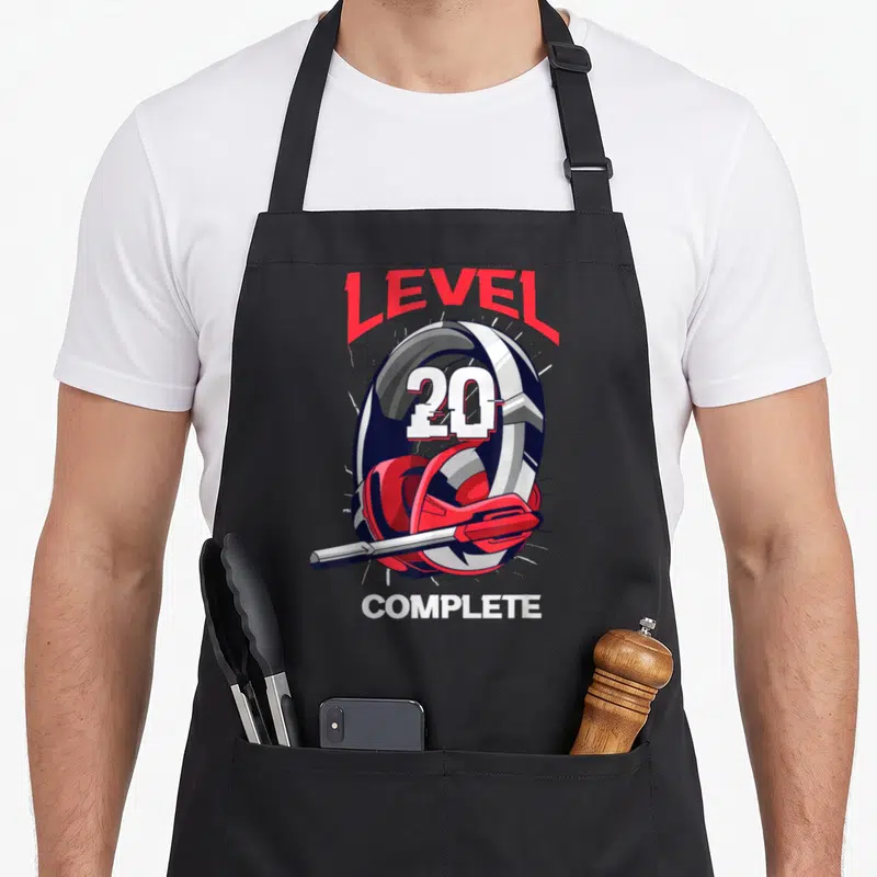 Tablier de cuisine noir avec illustration casque gaming et texte Level 20 Complete en rouge et blanc.
