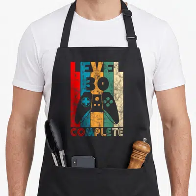 Tablier de cuisine noir avec illustration manette de jeu et texte Level 30 Complete sur fond de bandes colorées verticales.