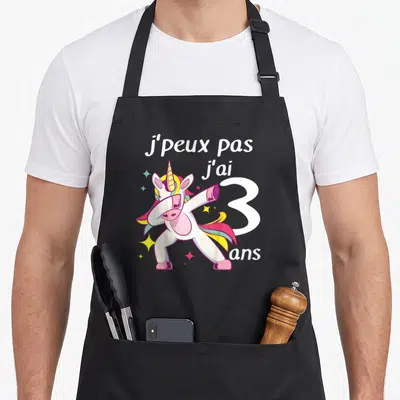 Tablier de cuisine noir avec une licorne colorée effectuant un mouvement de danse dab au centre du motif imprimé.