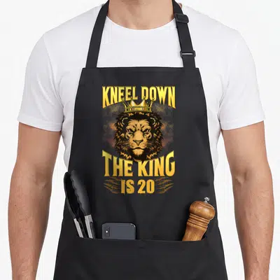 Tablier de cuisine noir avec tête de lion couronnée et texte doré Kneel Down The King Is 20.
