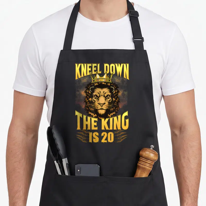 Tablier de cuisine noir avec tête de lion couronnée et texte doré Kneel Down The King Is 20.