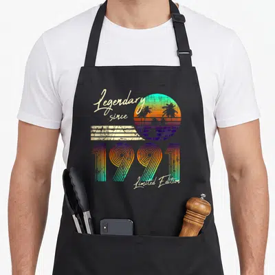 Tablier de cuisine noir avec motif rétro 1991, soleil coloré, palmiers et texte Legendary since Limited Edition en écriture cursive.