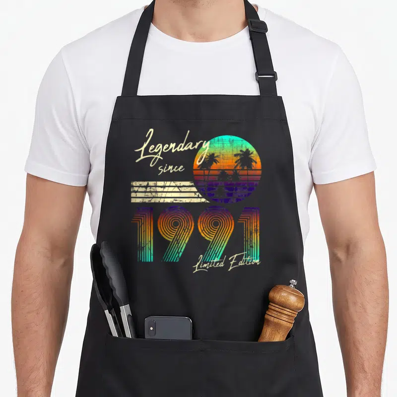 Tablier de cuisine noir avec motif rétro 1991, soleil coloré, palmiers et texte Legendary since Limited Edition en écriture cursive.