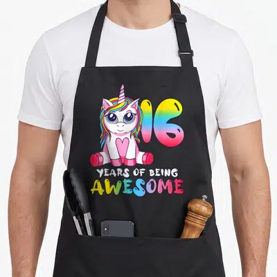 Tablier de cuisine noir avec licorne colorée, chiffre 16 arc-en-ciel et texte years of being « génial » en bas.