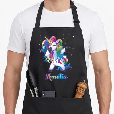 Tablier de cuisine noir avec une licorne aux cheveux arc-en-ciel effectuant un dab au-dessus du prénom Amélia en lettres colorées.