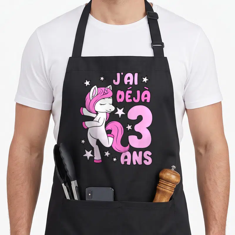 Tablier de cuisine noir avec licorne rose posant à côté du texte J'ai déjà 3 ans entouré d'étoiles blanches.