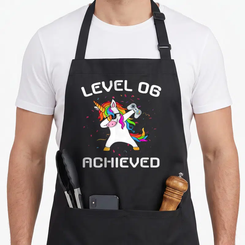 Tablier de cuisine noir avec une licorne arc-en-ciel en train de faire un dab tenant une manette de jeu vidéo.