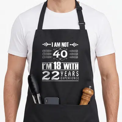 Tablier de cuisine noir avec texte blanc I AM NOT 40 I'M 18 WITH 22 YEARS EXPERIENCE et motifs décoratifs.