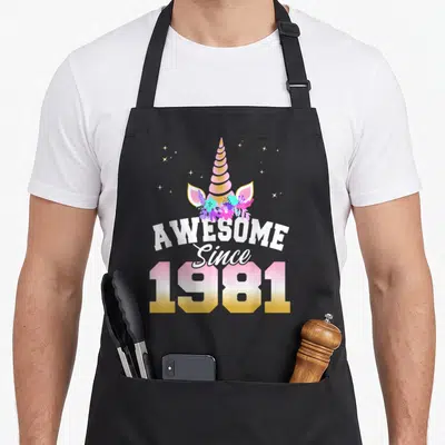 Tablier de cuisine noir avec illustration de licorne rose et texte Génial Depuis 1981 en dégradé doré.
