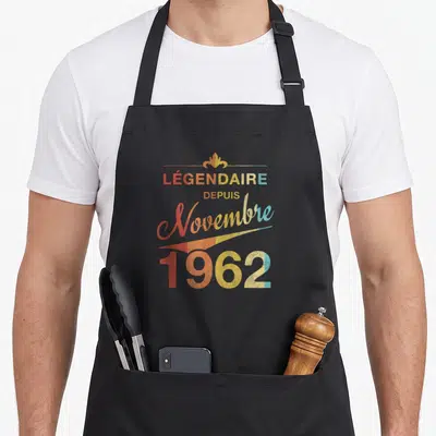 Tablier de cuisine noir avec inscription légendaire depuis novembre 1962 en lettres colorées style vintage et motif décoratif doré au sommet.