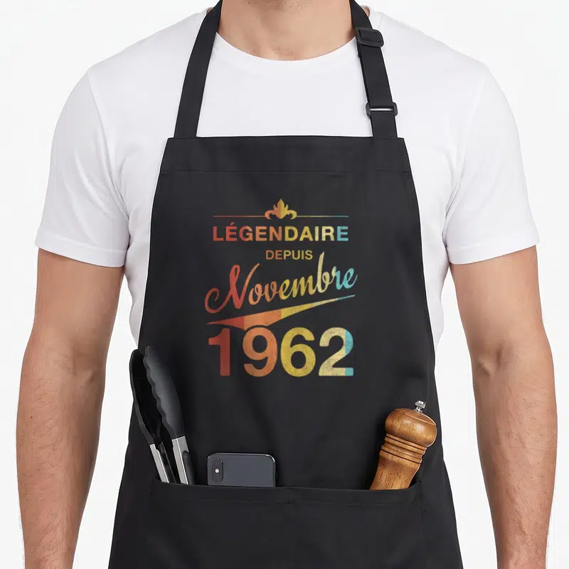 Tablier de cuisine noir avec inscription légendaire depuis novembre 1962 en lettres colorées style vintage et motif décoratif doré au sommet.