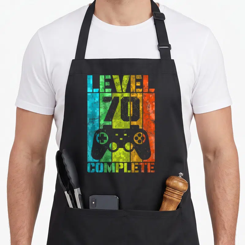 Tablier de cuisine noir avec motif pixelisé multicolore affichant Level 70 Complete et une silhouette de manette de jeu vidéo grise au centre.