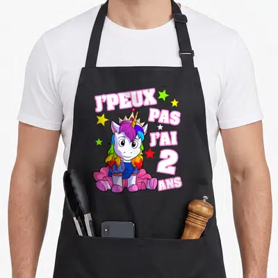 Tablier de cuisine noir avec licorne rose couronnée et texte J'peux pas j'ai 2 ans entouré d'étoiles colorées.