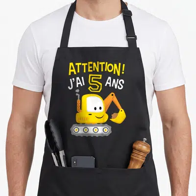 Tablier de cuisine noir avec une pelleteuse jaune souriante et le texte Attention j'ai 5 ans imprimé en lettres colorées.