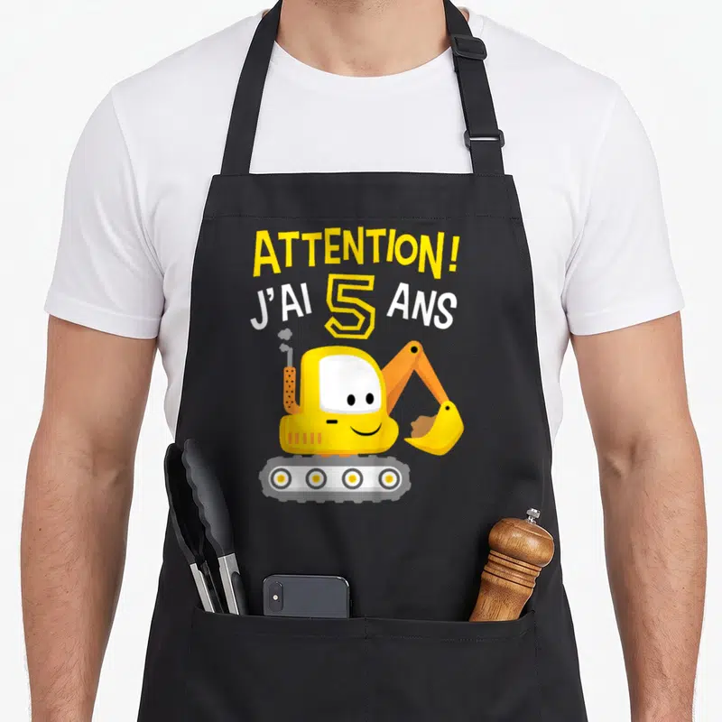 Tablier de cuisine noir avec une pelleteuse jaune souriante et le texte Attention j'ai 5 ans imprimé en lettres colorées.