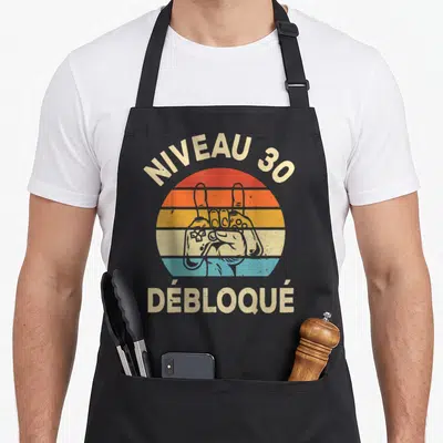 Tablier de cuisine noir avec une main tenant une manette devant un soleil arc-en-ciel et l'inscription Niveau 30 Débloqué.
