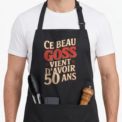Tablier de cuisine noir avec l'inscription Ce beau goss vient d'avoir 50 ans en lettres rouges et crème.