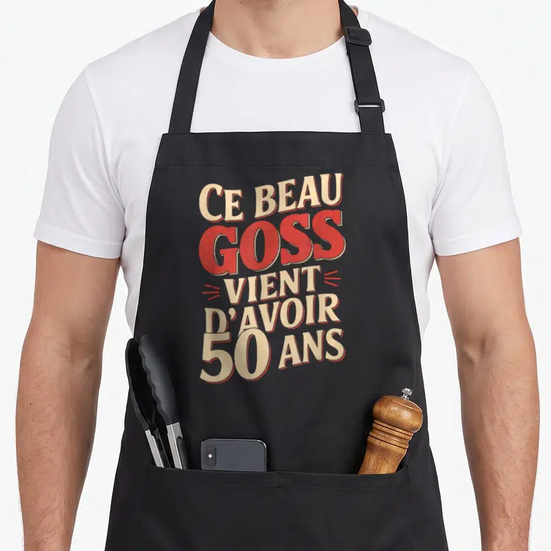 Tablier de cuisine noir avec l'inscription Ce beau goss vient d'avoir 50 ans en lettres rouges et crème.